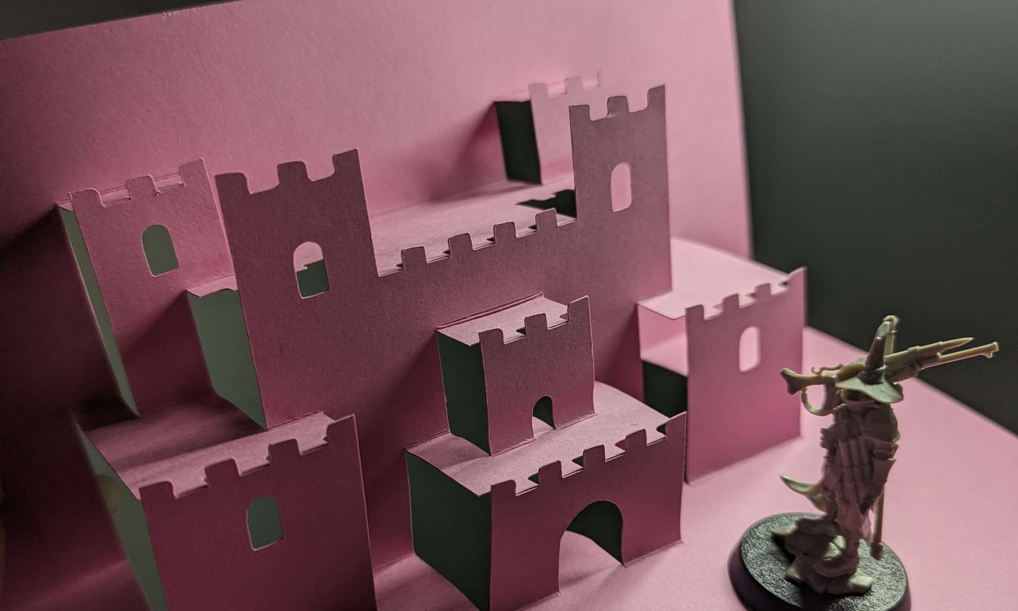 a figurine with a wizard hat and a gun stands next to a three-tiered pop up castle made by folding a single sheet of pink cardstock 90 degrees.